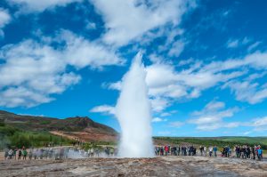 Tourists gathered around Strokkur geyser in the Haukadalur geothermal area of Iceland, watching it erupt powerfully against a backdrop of hills, greenery, and a bright blue sky with scattered clouds.
