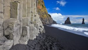 Columnar basalt formations on Reynisfjara black sand beach with Reynisdrangar sea stacks visible offshore, showcasing volcanic activity and coastal landforms for geography education.
