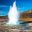 Eruption of Strokkur geyser in Iceland, shooting a column of water into the air against a backdrop of snow-capped mountains, illustrating geothermal activity for geography students.