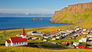 Panoramic view of the coastal village of Vík í Mýrdal in South Iceland, showing the iconic Reynisdrangar sea stacks, basalt cliffs, and red-roofed church — a picturesque stop on geography school trips focused on coastal erosion and volcanic landscapes.