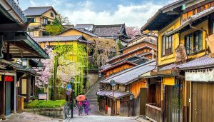 Traditional Kyoto old town street with historic wooden buildings, blooming cherry blossom trees, and two women in kimonos walking with an oriental umbrella during springtime, perfect for cultural school trips in Japan.