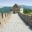 A scenic view along the Great Wall of China, showing the historic stone pathway stretching into the distance with mountainous green terrain in the background. Traditional Chinese watchtowers line the wall, and a few tourists are visible walking along the path.