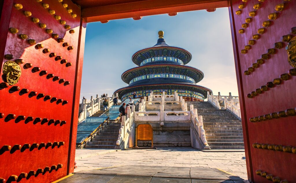 The Temple of Heaven in Beijing, China, viewed through an open red gate adorned with golden studs. The circular, multi-tiered blue and gold temple rises behind a broad staircase, with visitors ascending the steps under a clear sky.