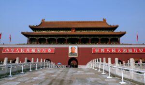 The Tiananmen Gate in Beijing, China, featuring traditional Chinese architecture with a large portrait of Chairman Mao Zedong above the central entrance. Red banners with Chinese characters are displayed on either side, and visitors walk along the white stone pathway leading to the entrance.